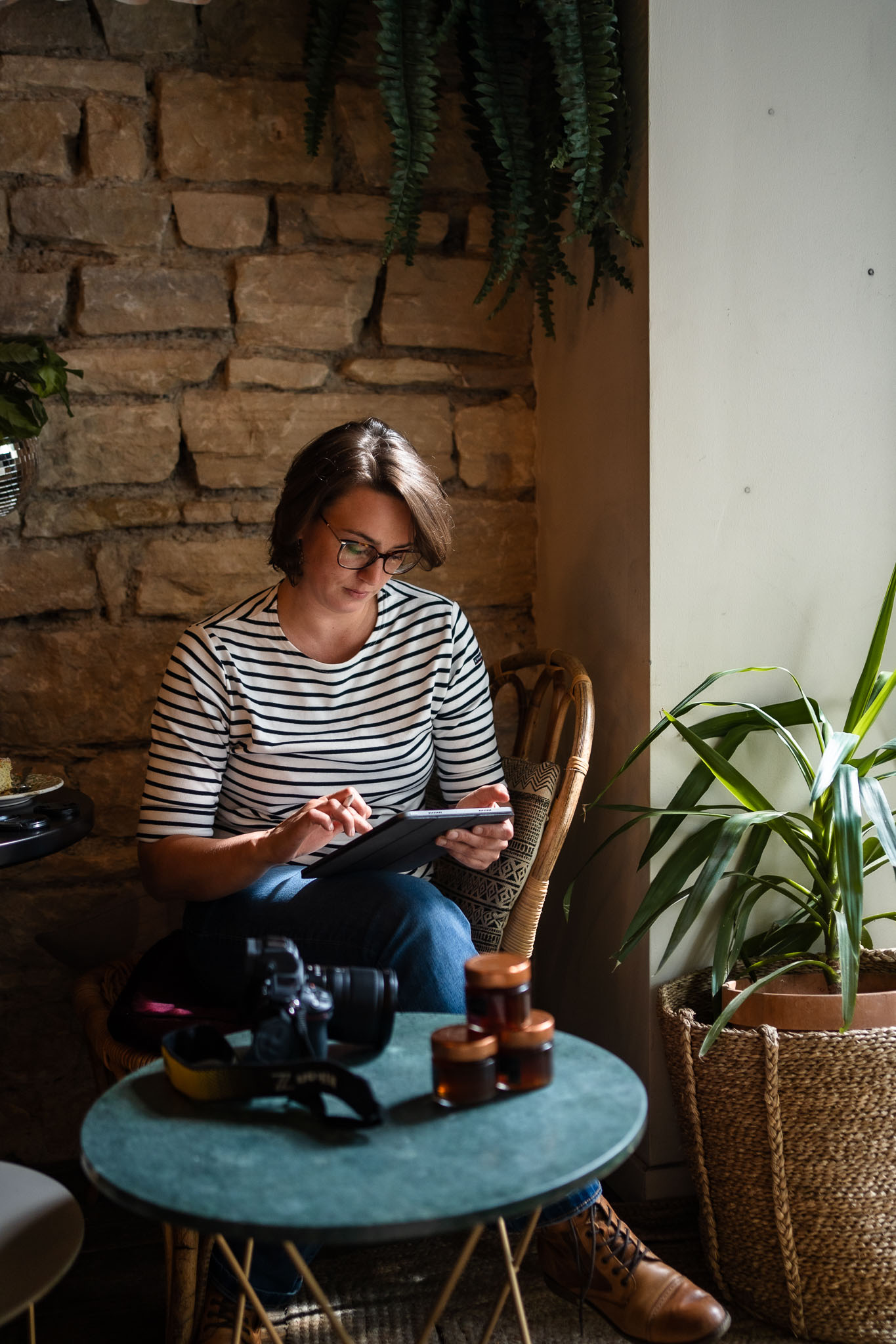 Une femme portant des lunettes et une marinière, assise sur une chaise en osier, utilise une tablette. Elle est entourée de plantes vertes et d'une table basse avec un appareil photo et des accessoires