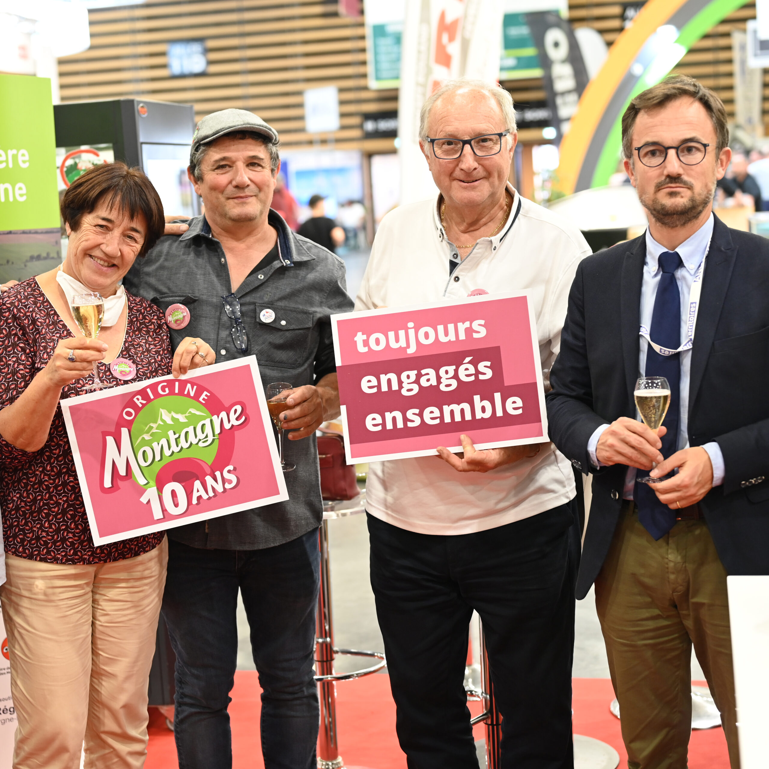 Quatre personnes posant ensemble lors d'un événement, tenant des panneaux célébrant les 10 ans d'Origine Montagne avec le slogan 'toujours engagés ensemble', illustrant une campagne de communication axée sur l'engagement continu et la célébration communautaire.