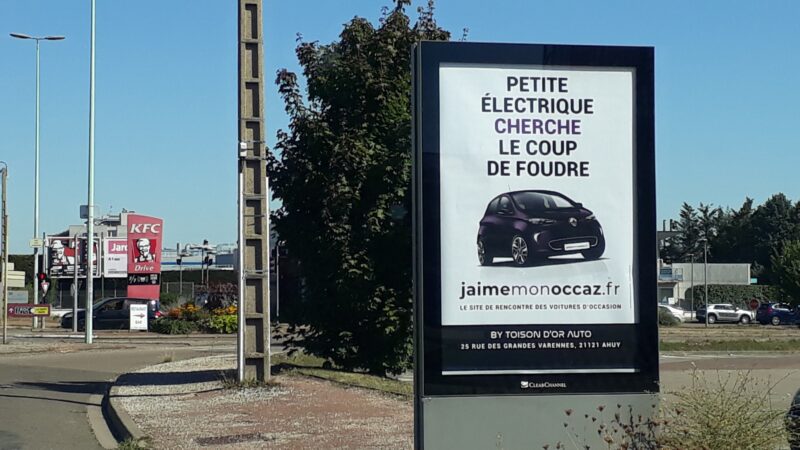branding communication Panneau publicitaire en bord de route pour un site de voitures d'occasion, mettant en avant la recherche de véhicules électriques avec le slogan "Cherche le coup de foudre"